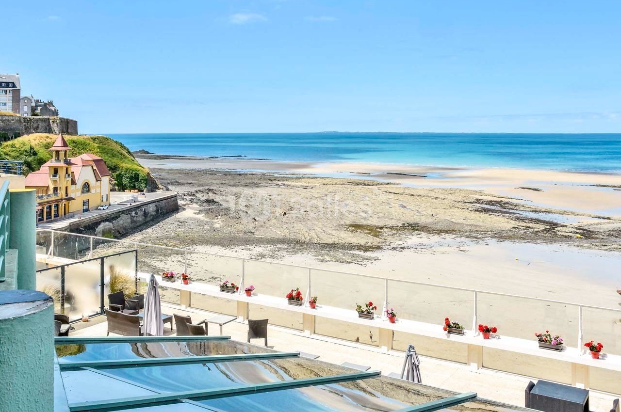 Vue sur une plage de sable à marée basse avec des bâtiments en bord de mer et une terrasse ornée de fleurs.