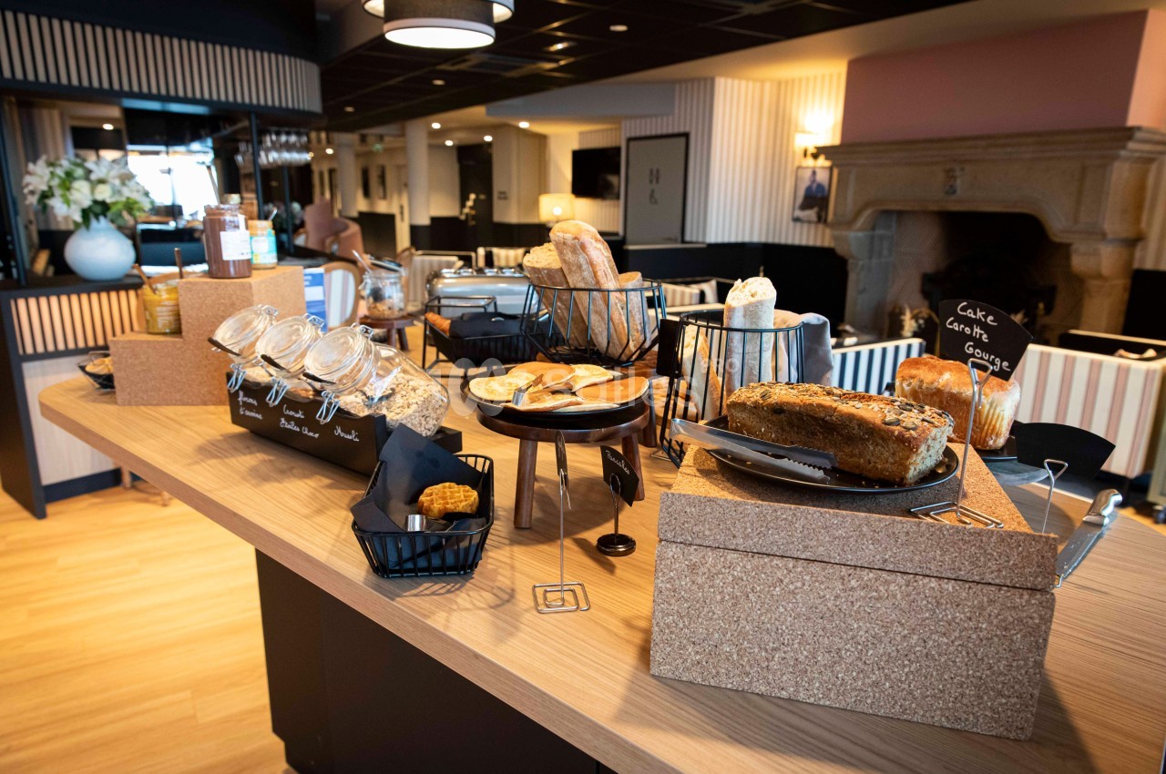 Buffet de petit-déjeuner avec pains, céréales, gâteaux et condiments disposés sur une table en bois dans une salle lumineuse.