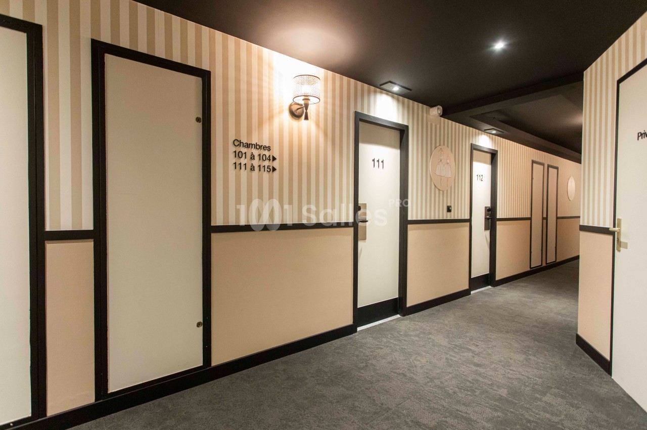 Couloir d'hôtel avec plusieurs portes numérotées, éclairage mural et moquette grise.