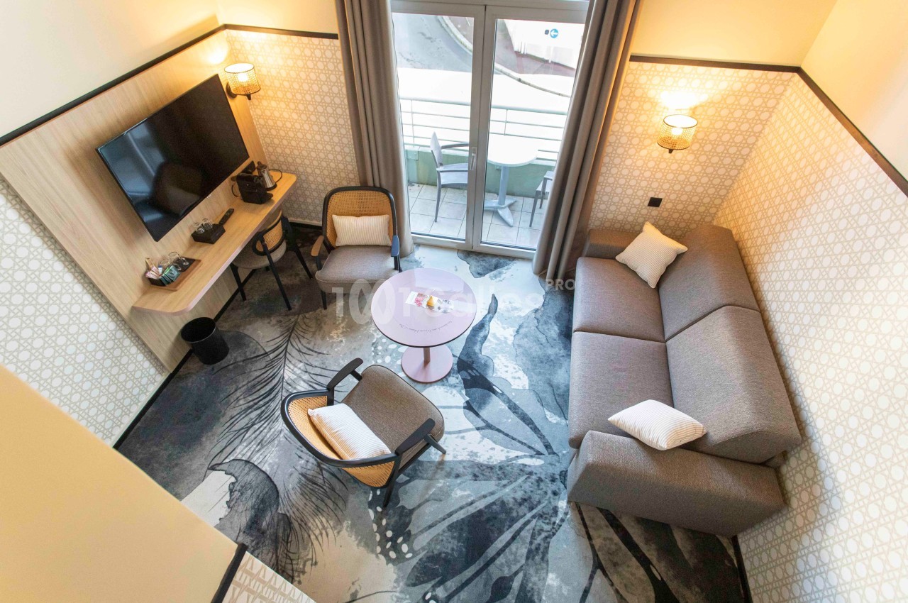 Salon moderne avec canapé, fauteuils, table basse, télévision murale et accès à un balcon avec mobilier extérieur.