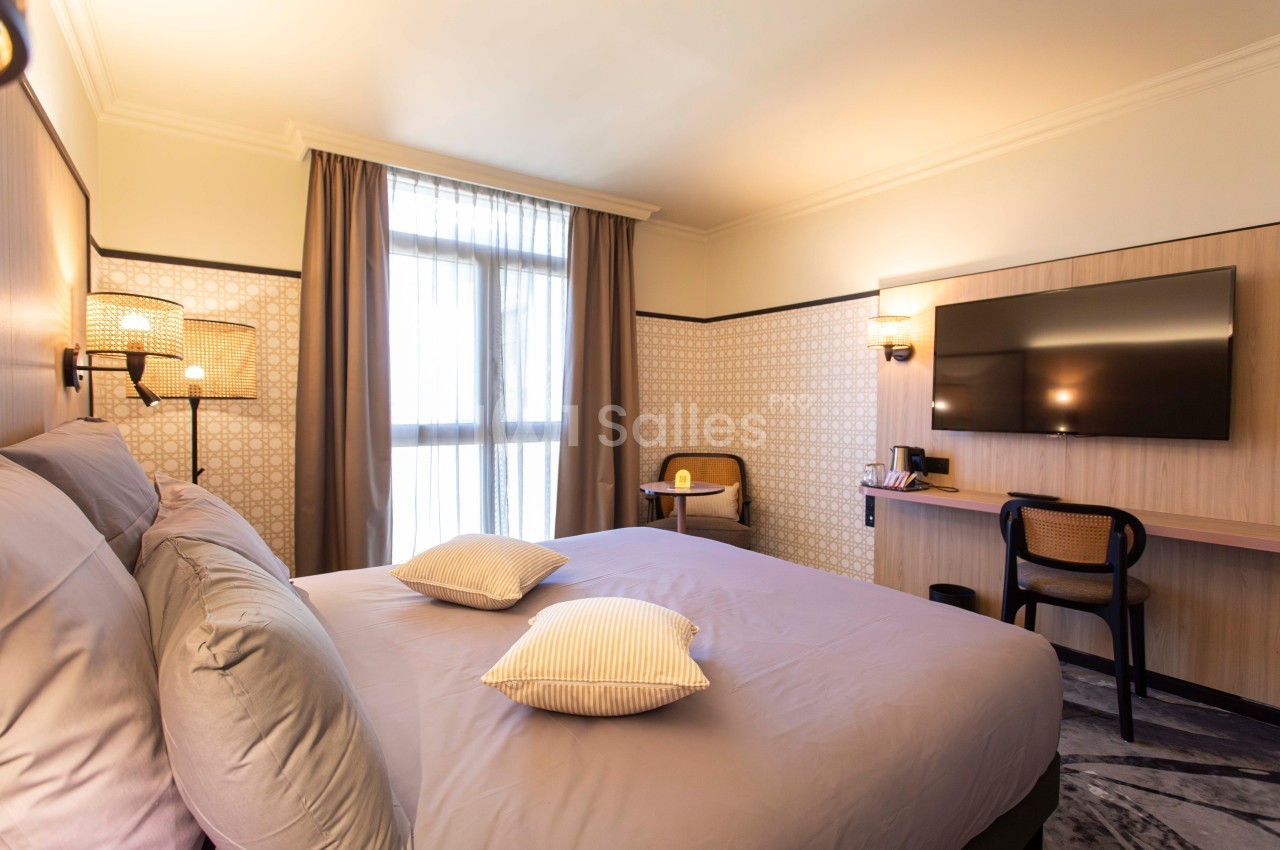 Chambre d'hôtel lumineuse avec lit double, télévision murale, bureau, chaise et grande fenêtre avec rideaux.