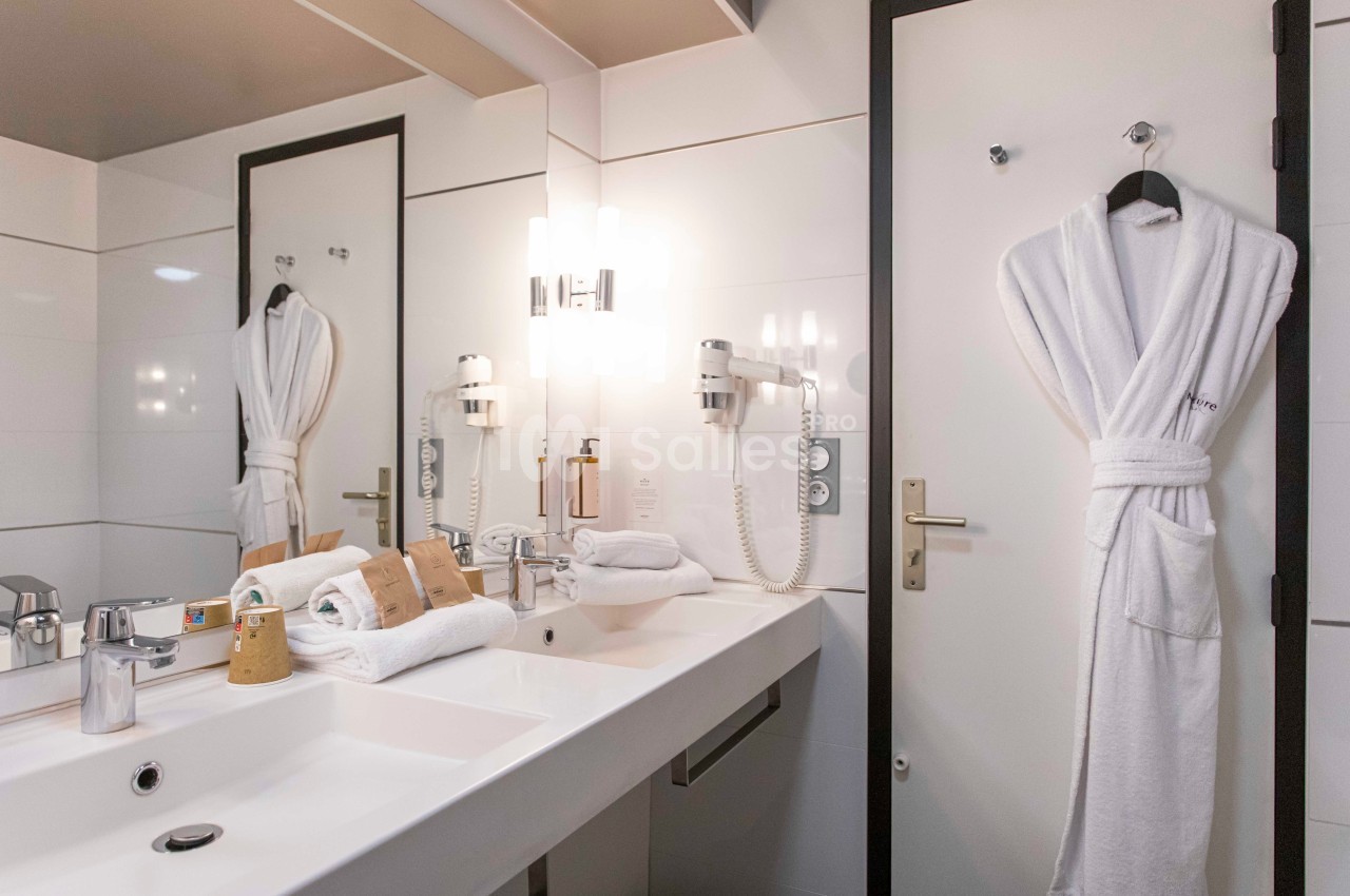 Salle de bain moderne avec double vasque, miroir éclairé, sèche-cheveux, peignoir suspendu et articles de toilette.