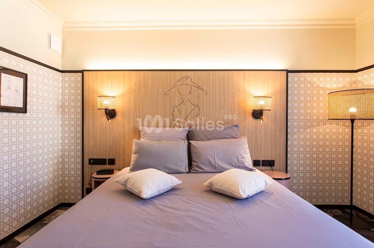 Lit double avec draps violets, deux lampes murales et une décoration murale en bois dans une chambre lumineuse.