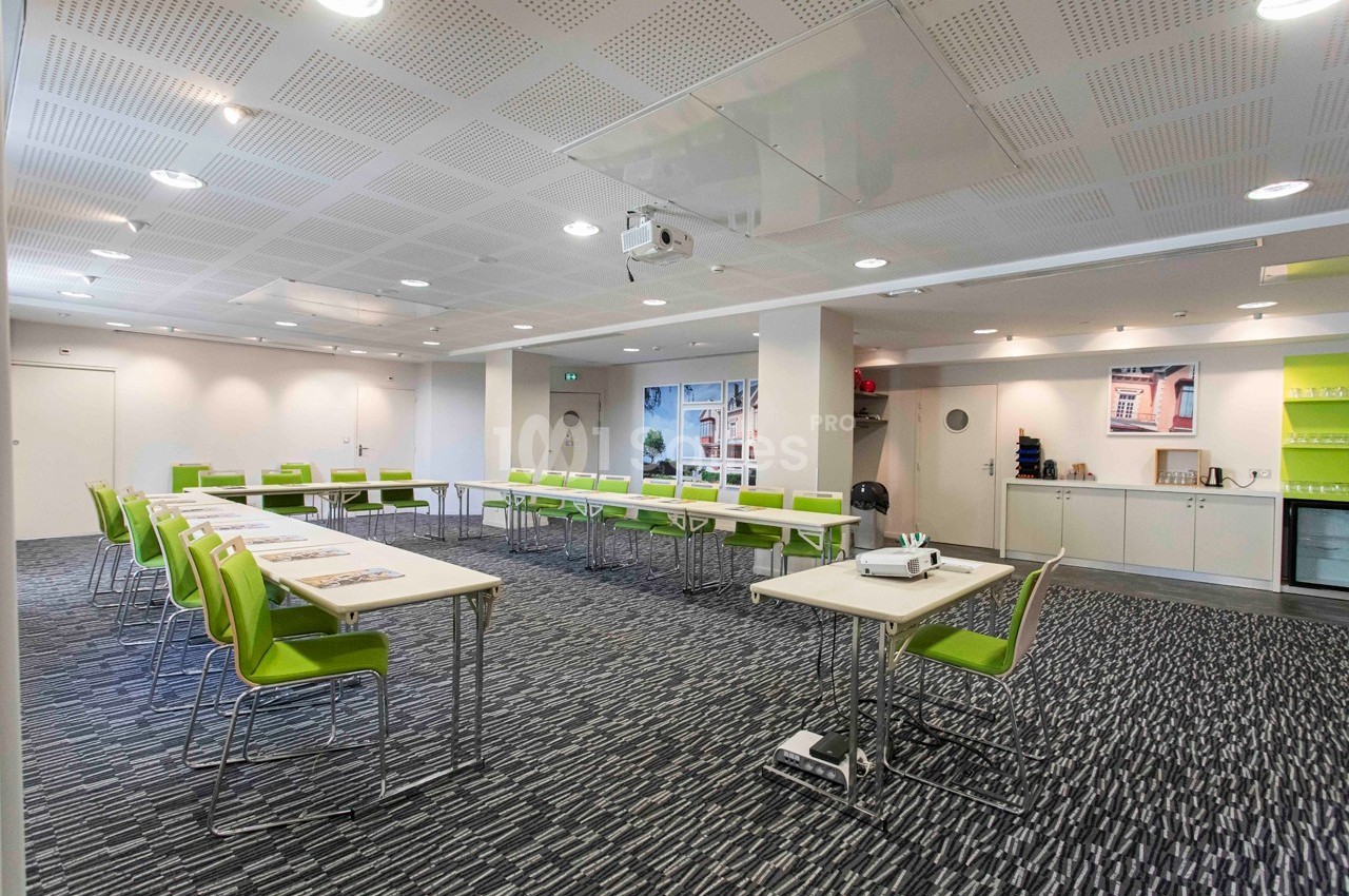 Salle de réunion lumineuse avec tables en U, chaises vertes, vidéoprojecteur et étagères en arrière-plan.
