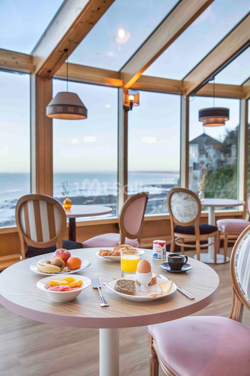 Petit-déjeuner servi sur une table ronde dans une salle lumineuse avec vue sur la mer à travers de grandes baies vitrées.