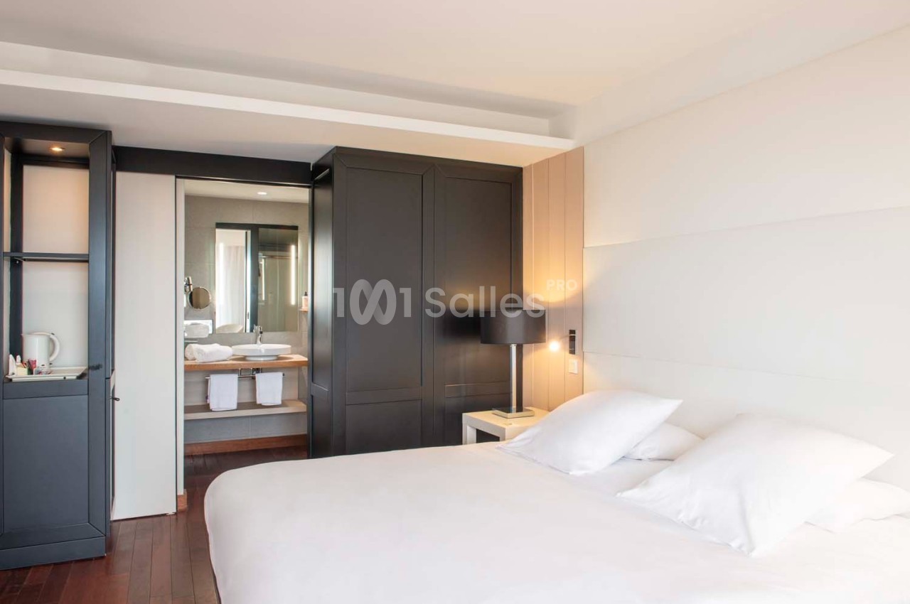 Chambre lumineuse avec lit double, armoire noire, table de chevet et vue sur une salle de bain moderne en arrière-plan.