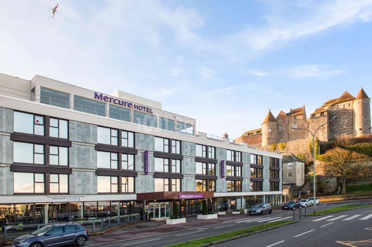 Façade d'un hôtel moderne avec un château en arrière-plan, situé près d'une route et sous un ciel dégagé.