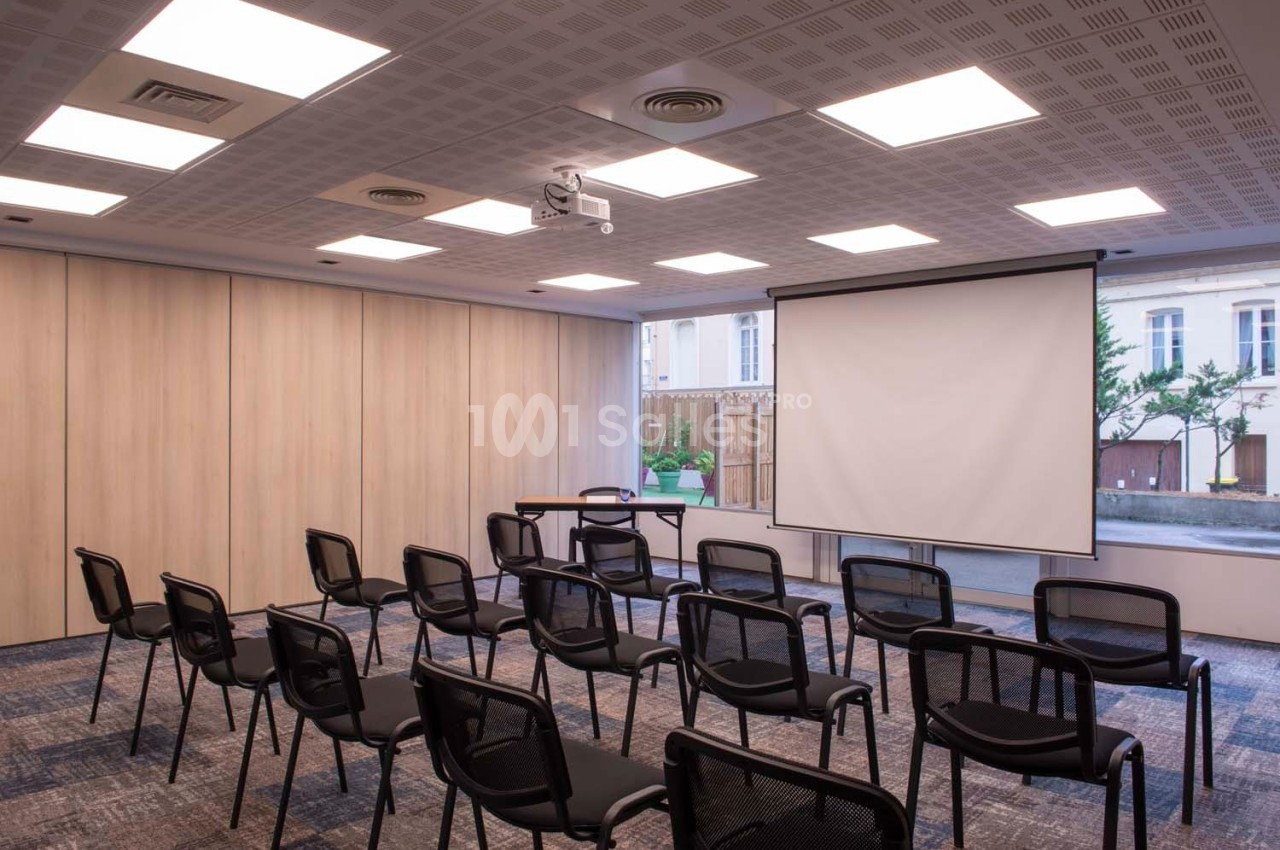 Salle de réunion avec chaises alignées, écran de projection déployé et lumière naturelle entrant par une baie vitrée.