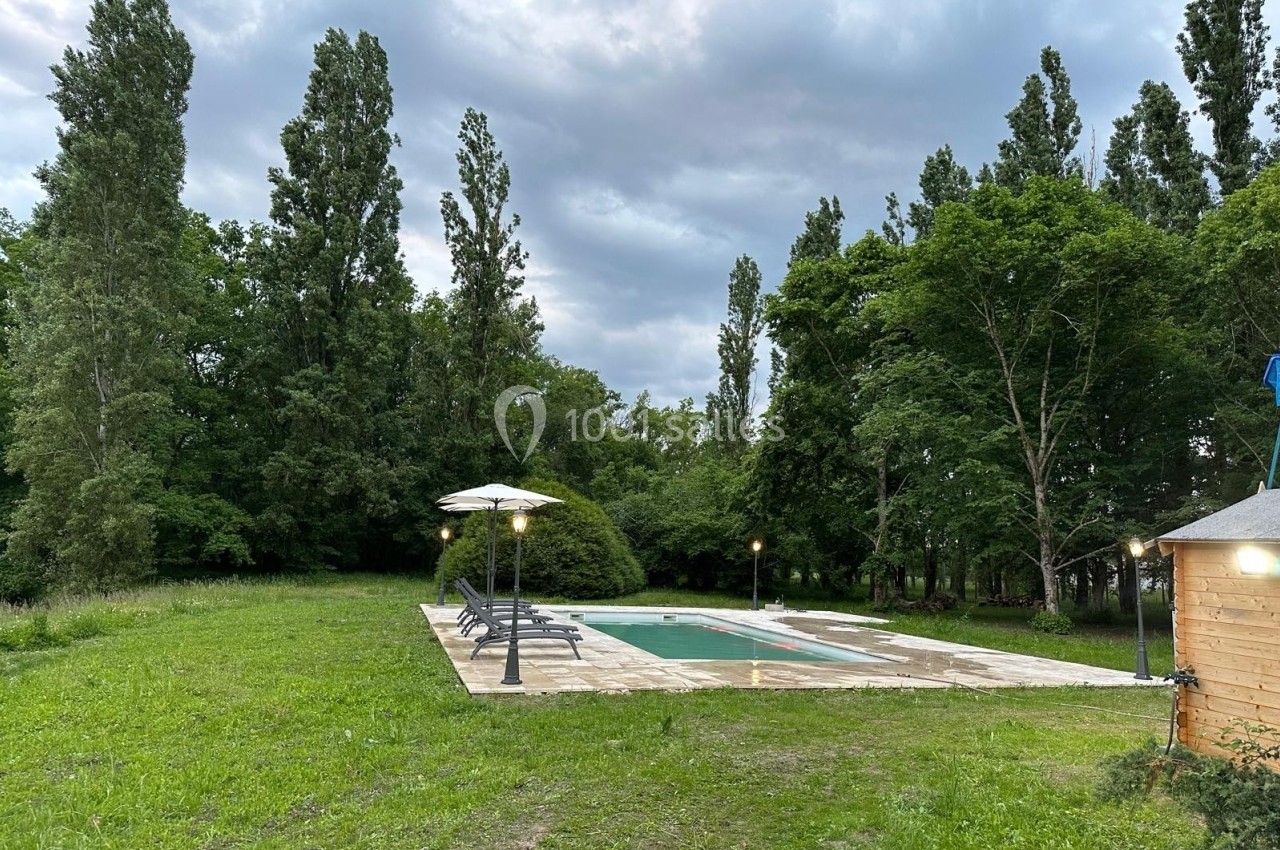 Piscine extérieure entourée de chaises longues, parasols et pelouse, bordée d'arbres sous un ciel nuageux.
