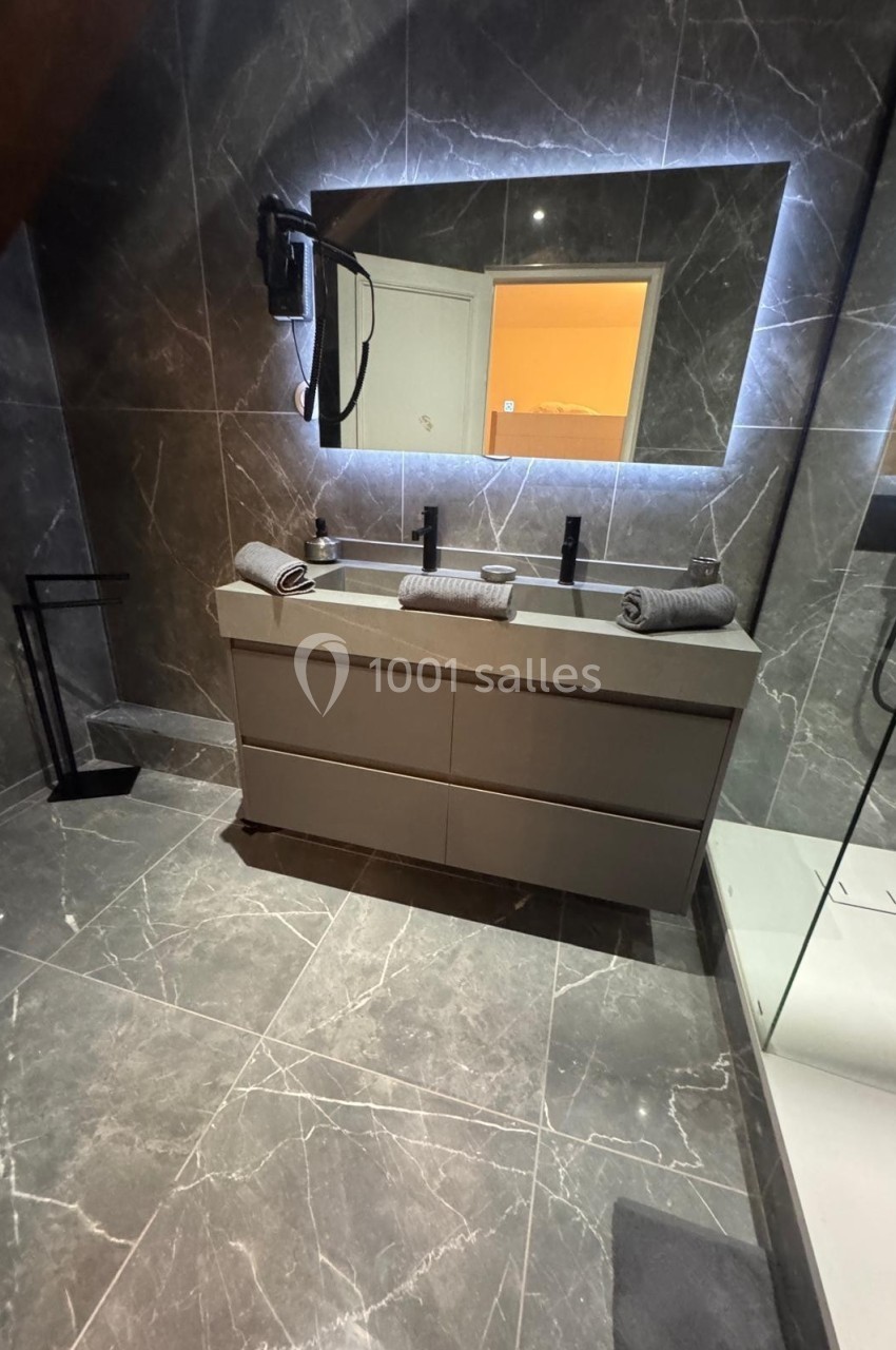 Salle de bain moderne avec double vasque, miroir rétroéclairé, douche vitrée et carrelage effet marbre.