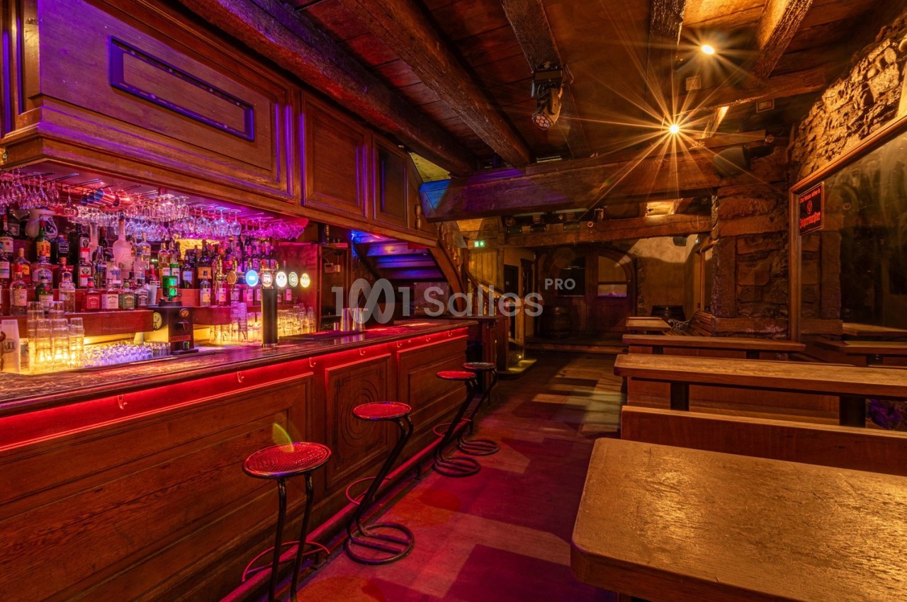 Bar en bois avec comptoir éclairé, tabourets hauts, étagères de bouteilles et tables en bois dans une ambiance chaleureuse.