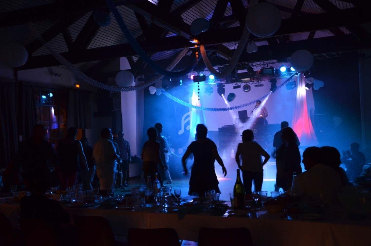 Des personnes dansent dans une salle sombre éclairée par des lumières colorées, avec des tables dressées au premier plan.
