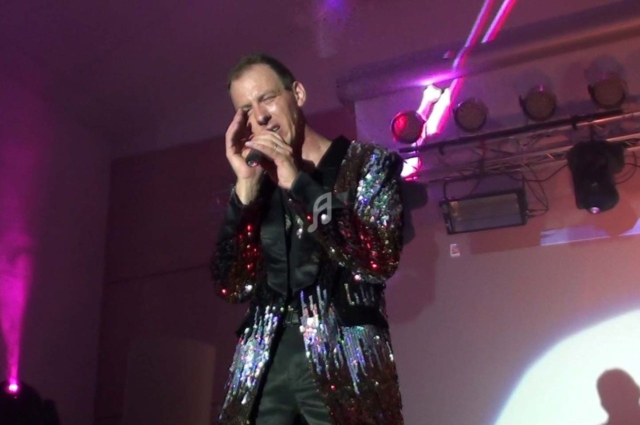 Homme chantant sur scène, vêtu d'une veste à paillettes, éclairé par des projecteurs colorés.
