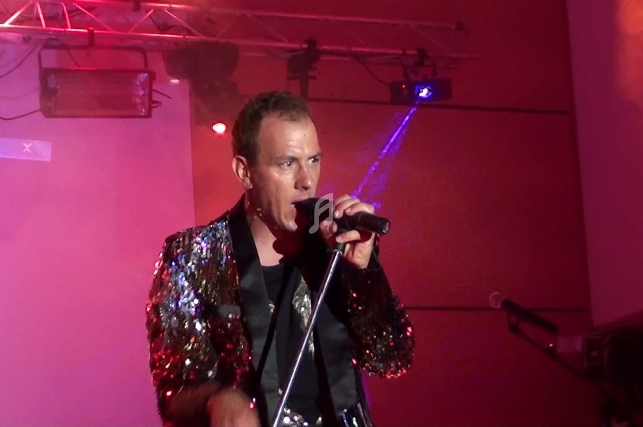 Un chanteur en veste à paillettes interprète sur scène, éclairé par des projecteurs rouges et violets.