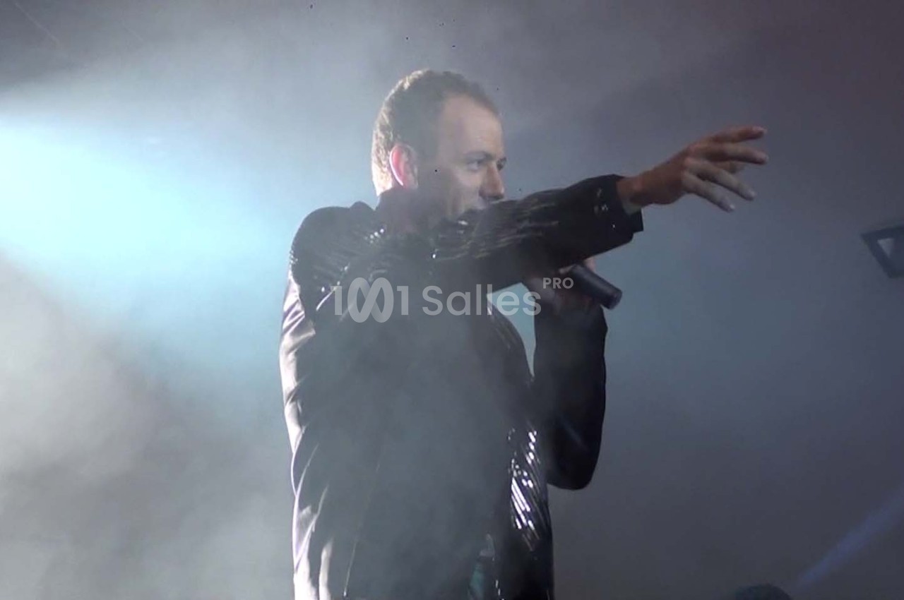 Un chanteur en veste noire tient un micro et pointe du doigt sur une scène éclairée par des projecteurs.
