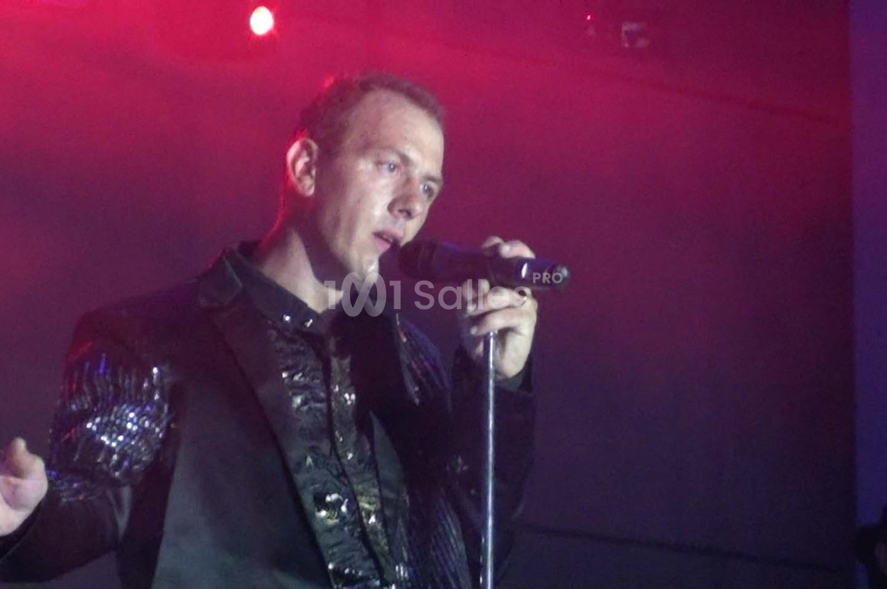 Un homme chante sur scène avec un micro, éclairé par une lumière rouge dans une ambiance de concert.