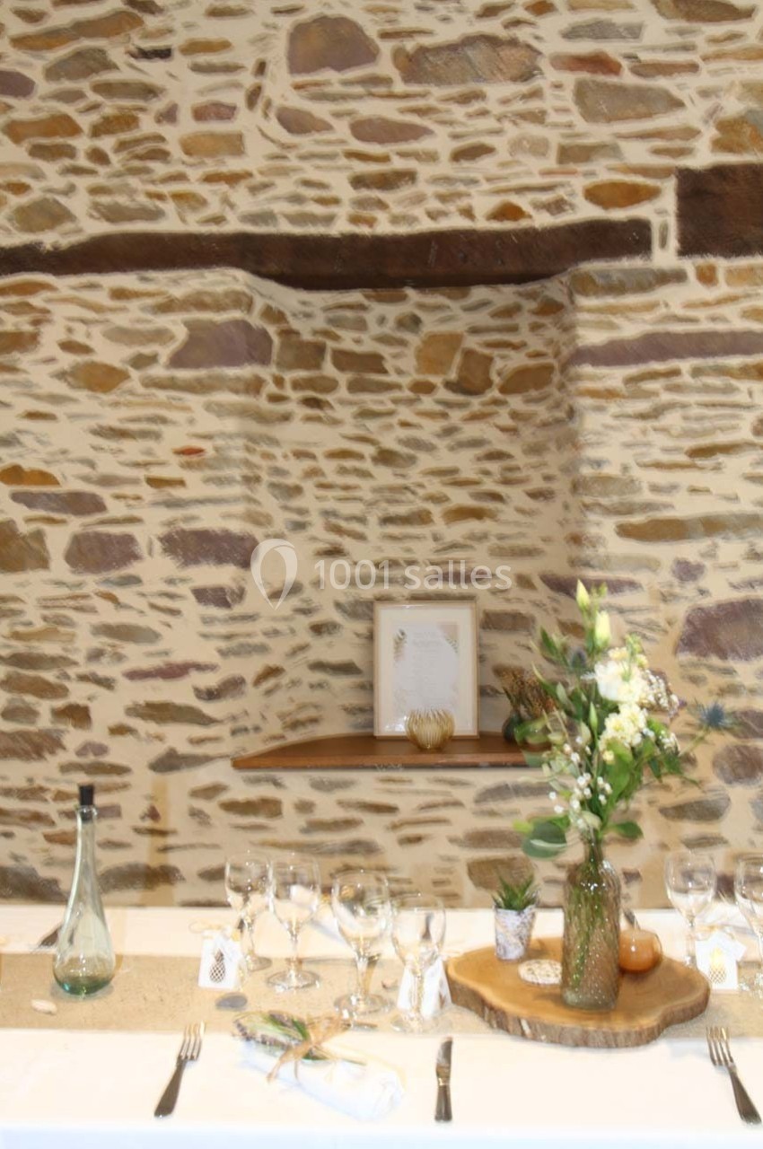 Table dressée avec des verres, couverts et décoration florale, devant un mur en pierre apparente.