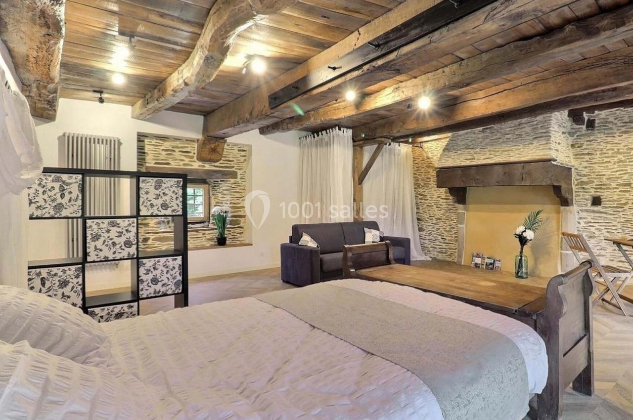 Chambre rustique avec poutres apparentes, lit double, coin salon, cheminée en pierre et décoration sobre.