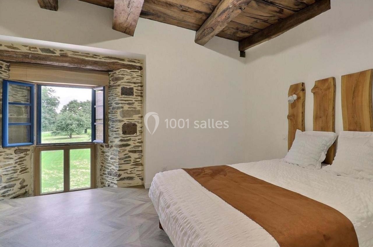 Chambre lumineuse avec lit double, murs en pierre, plafond en bois et fenêtre ouverte sur un paysage verdoyant.