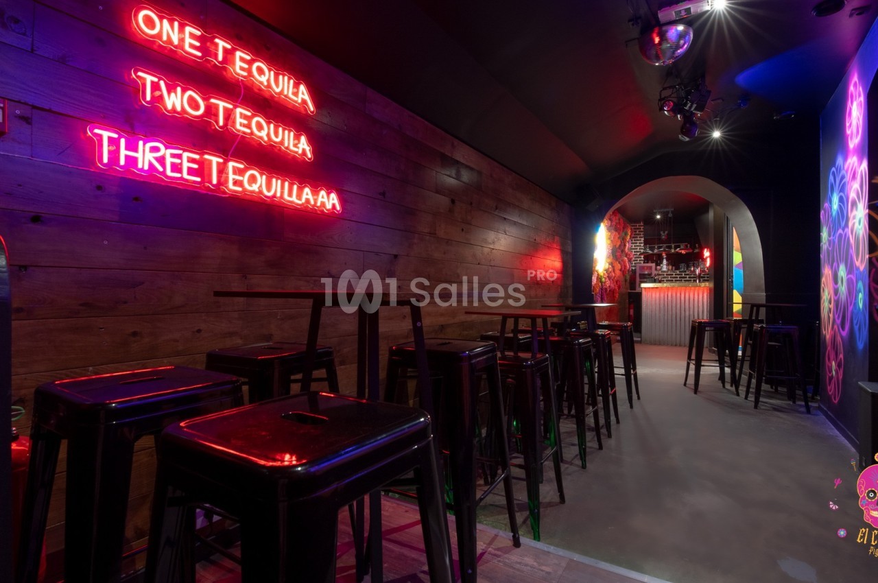Salle de bar avec murs en bois, néons roses, tabourets hauts et comptoir éclairé au fond.