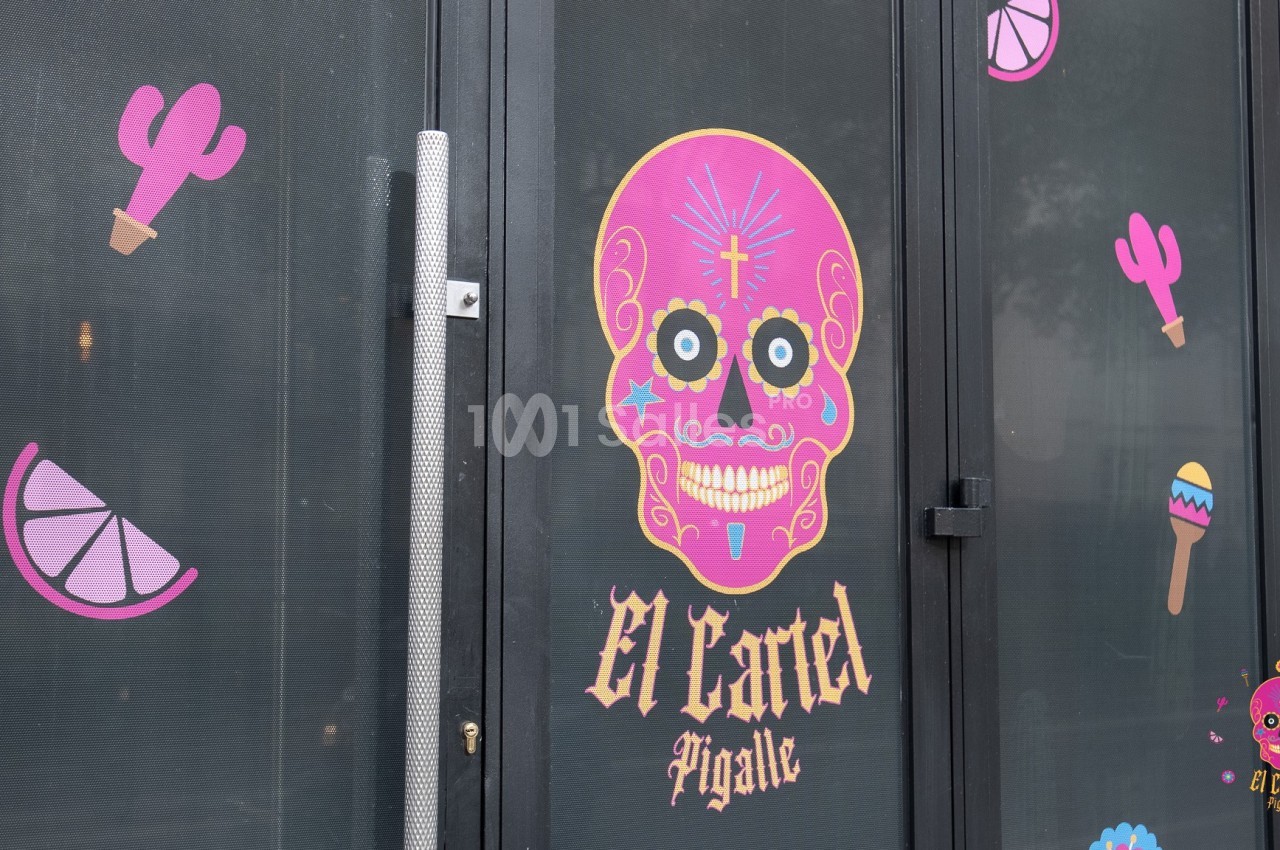 Autocollants colorés de motifs mexicains sur une porte vitrée, avec un crâne rose et l'inscription ’El Cartel Pigalle’.