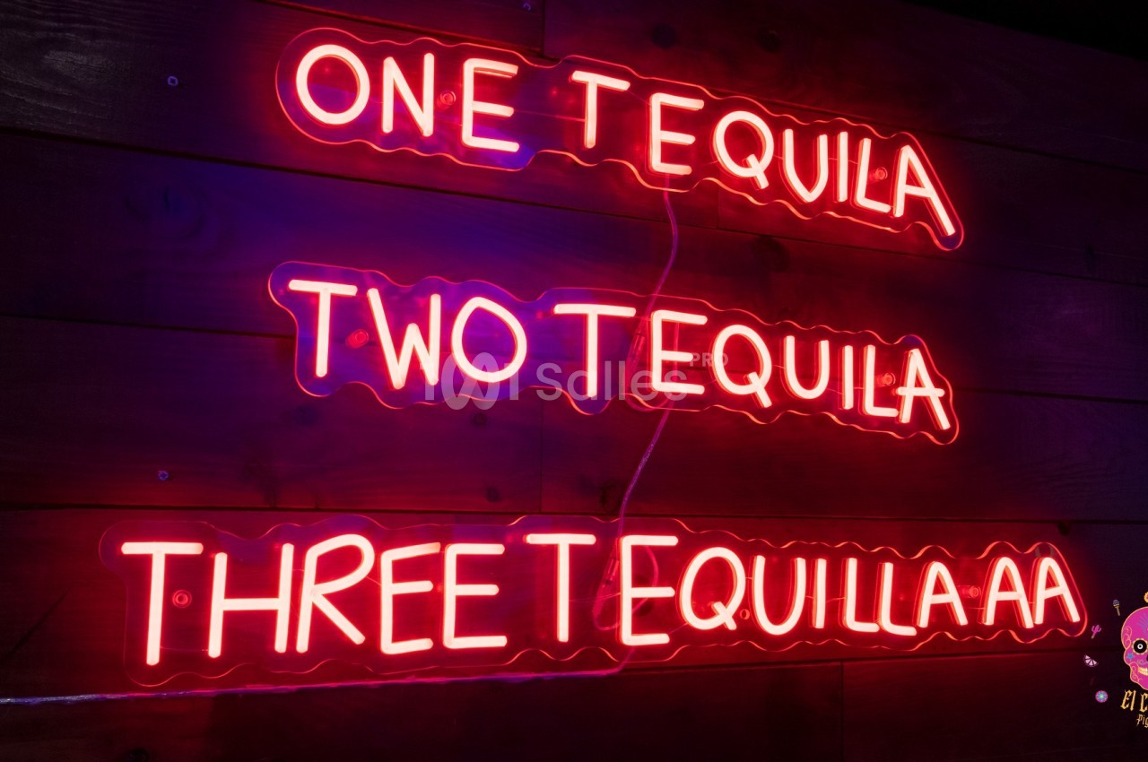 Enseigne lumineuse rose affichant ’One Tequila, Two Tequila, Three Tequilaaa’ sur un mur en bois sombre.