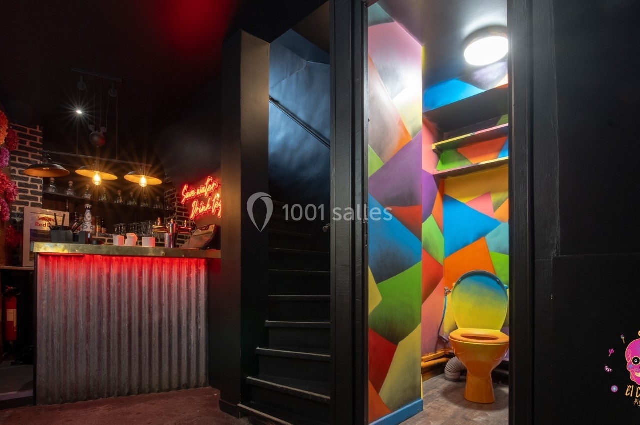 Bar avec comptoir éclairé, mur fleuri à gauche, escalier central et toilettes colorées à droite.