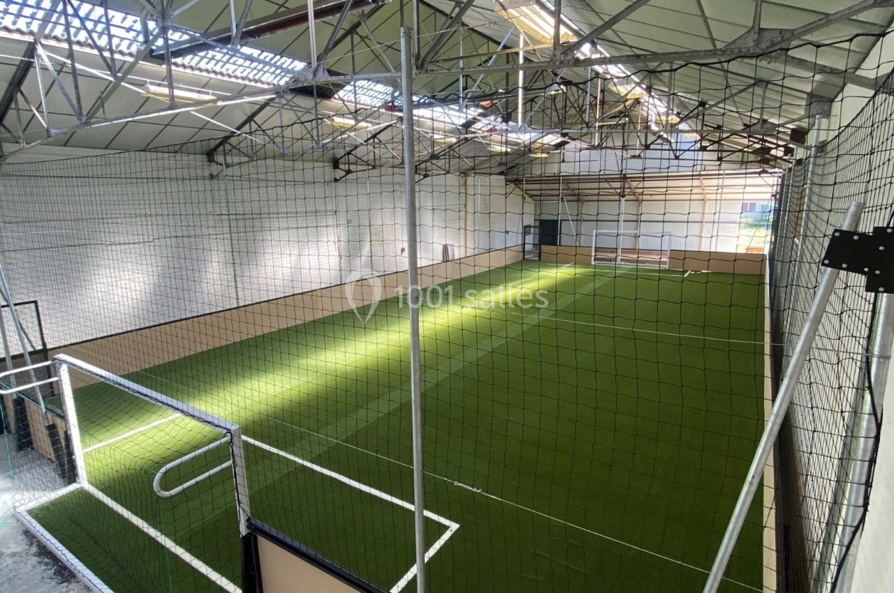 Terrain de sport intérieur avec pelouse synthétique, entouré de filets et situé sous une structure métallique.