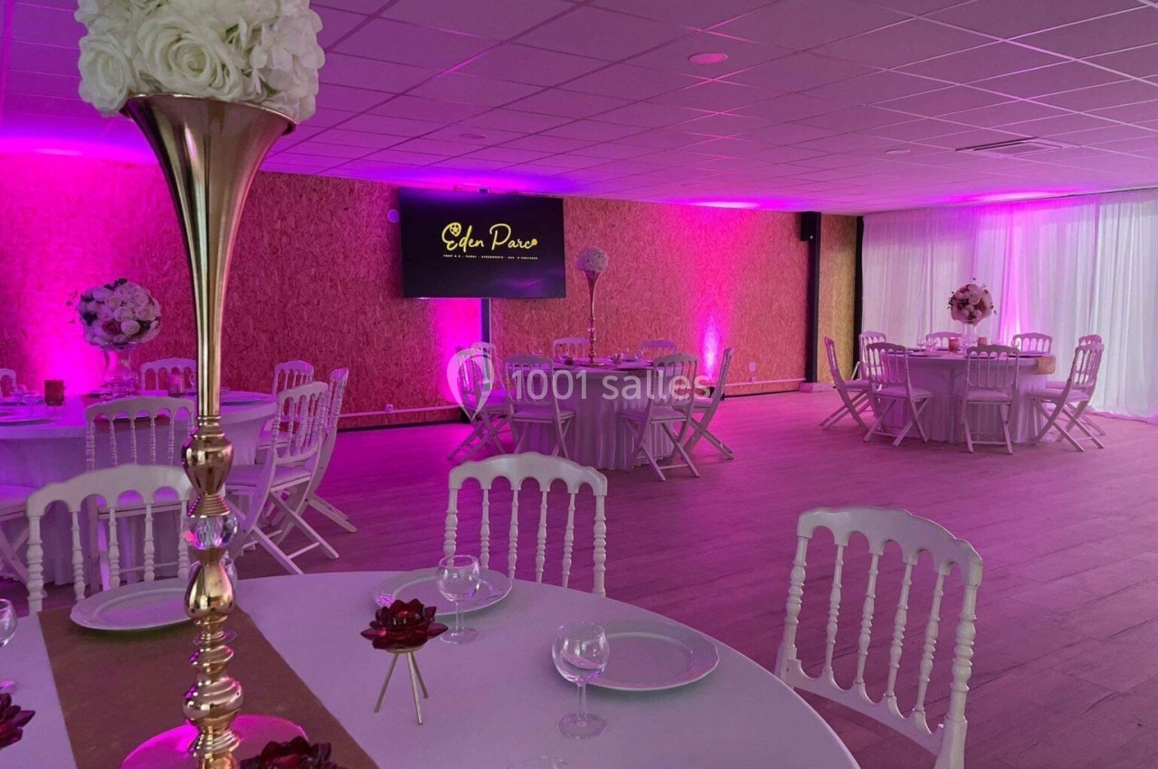 Salle de réception décorée avec des tables rondes, nappes blanches, chaises élégantes et éclairage rose tamisé.