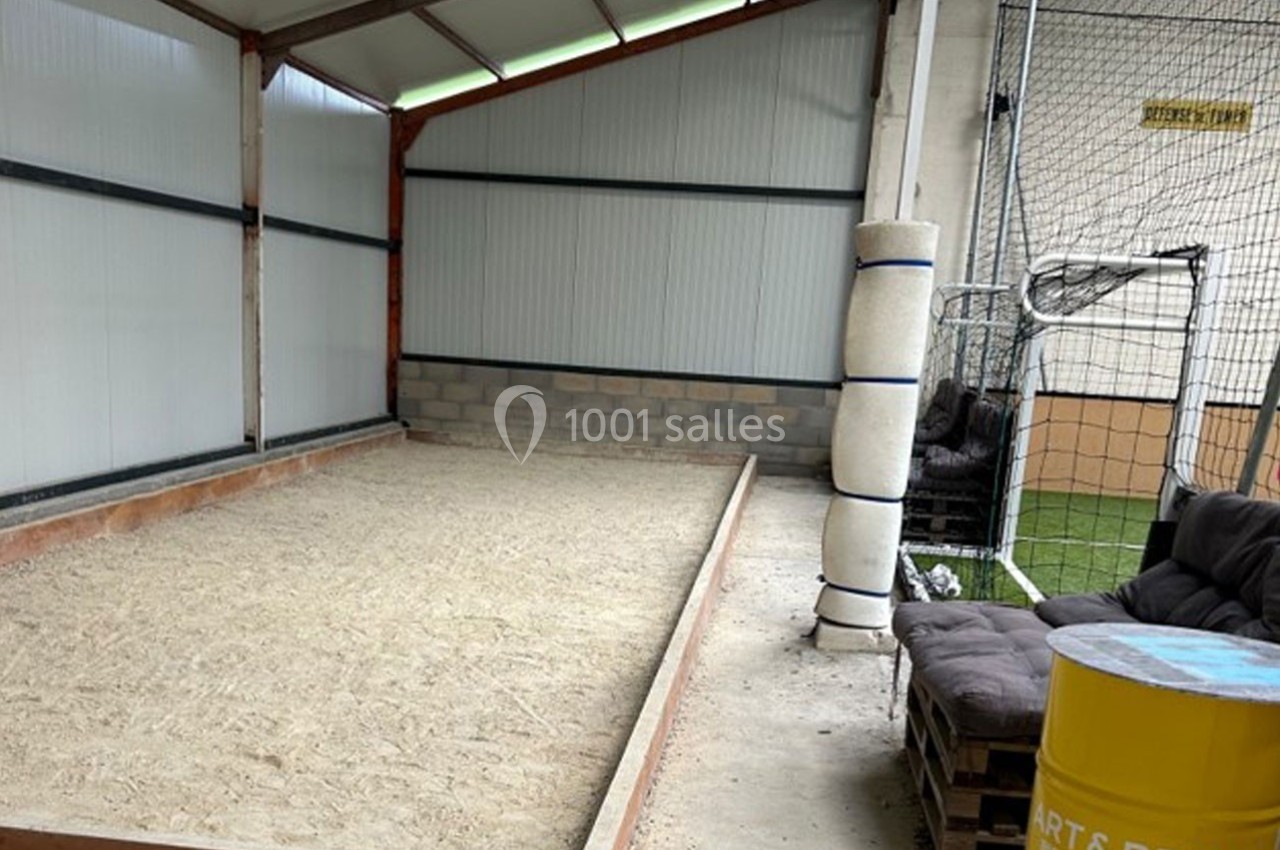 Terrain de pétanque intérieur avec sol en sable, encadré de murs en tôle et briques, à côté d'un espace détente.