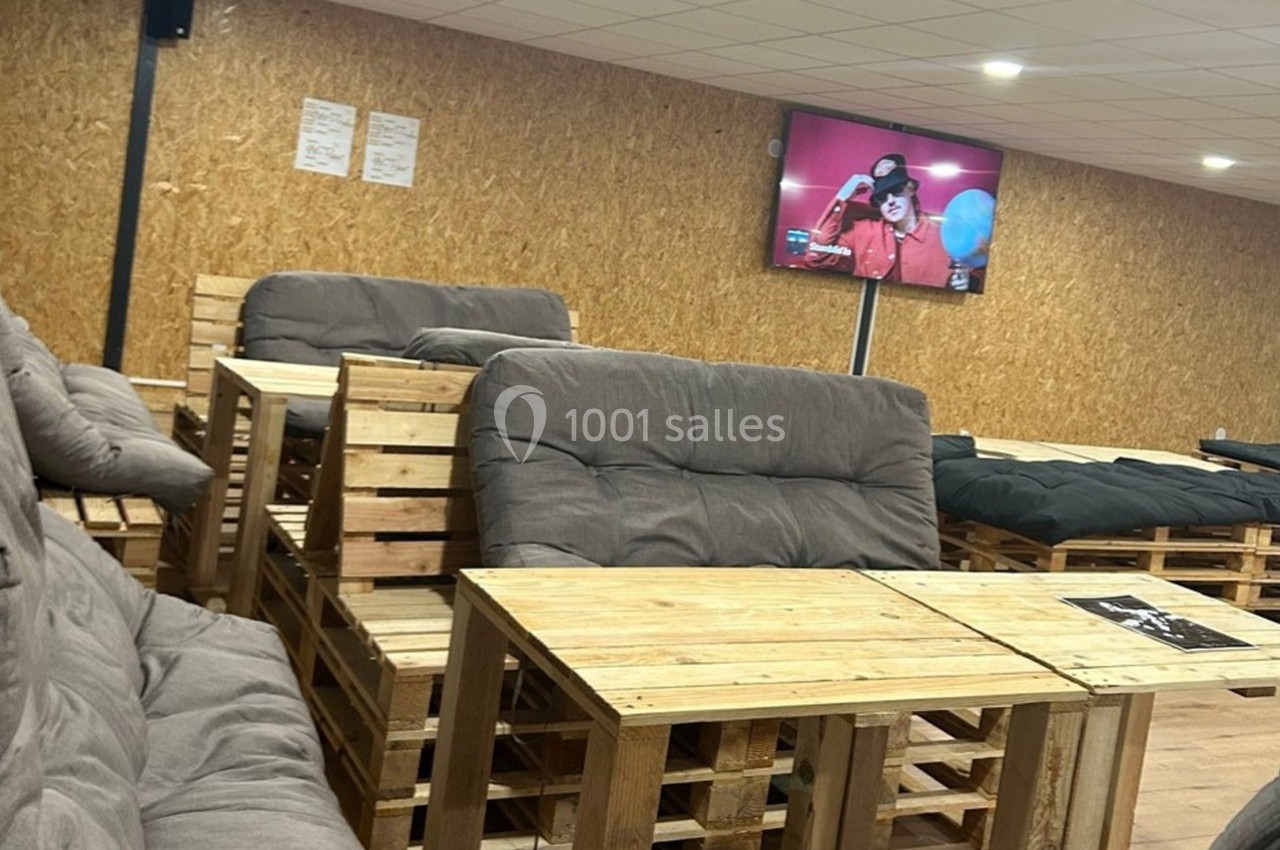 Salle aménagée avec des meubles en palettes, canapés gris et écran mural diffusant une vidéo.