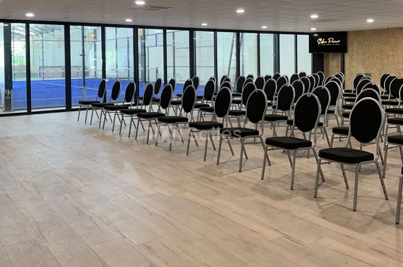 Salle de conférence lumineuse avec des rangées de chaises noires alignées face à de grandes baies vitrées.