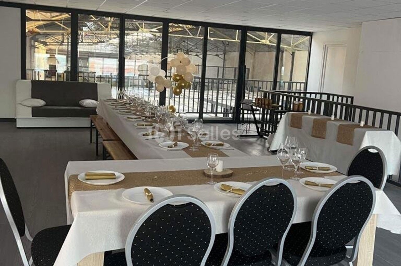 Salle de réception aménagée avec une longue table dressée, nappes blanches, décorations dorées et chaises noires.