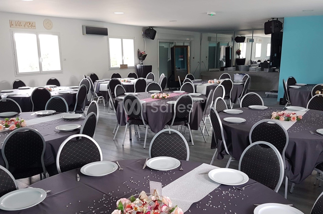 Salle de réception décorée avec des tables rondes, nappes grises, assiettes blanches et arrangements floraux.
