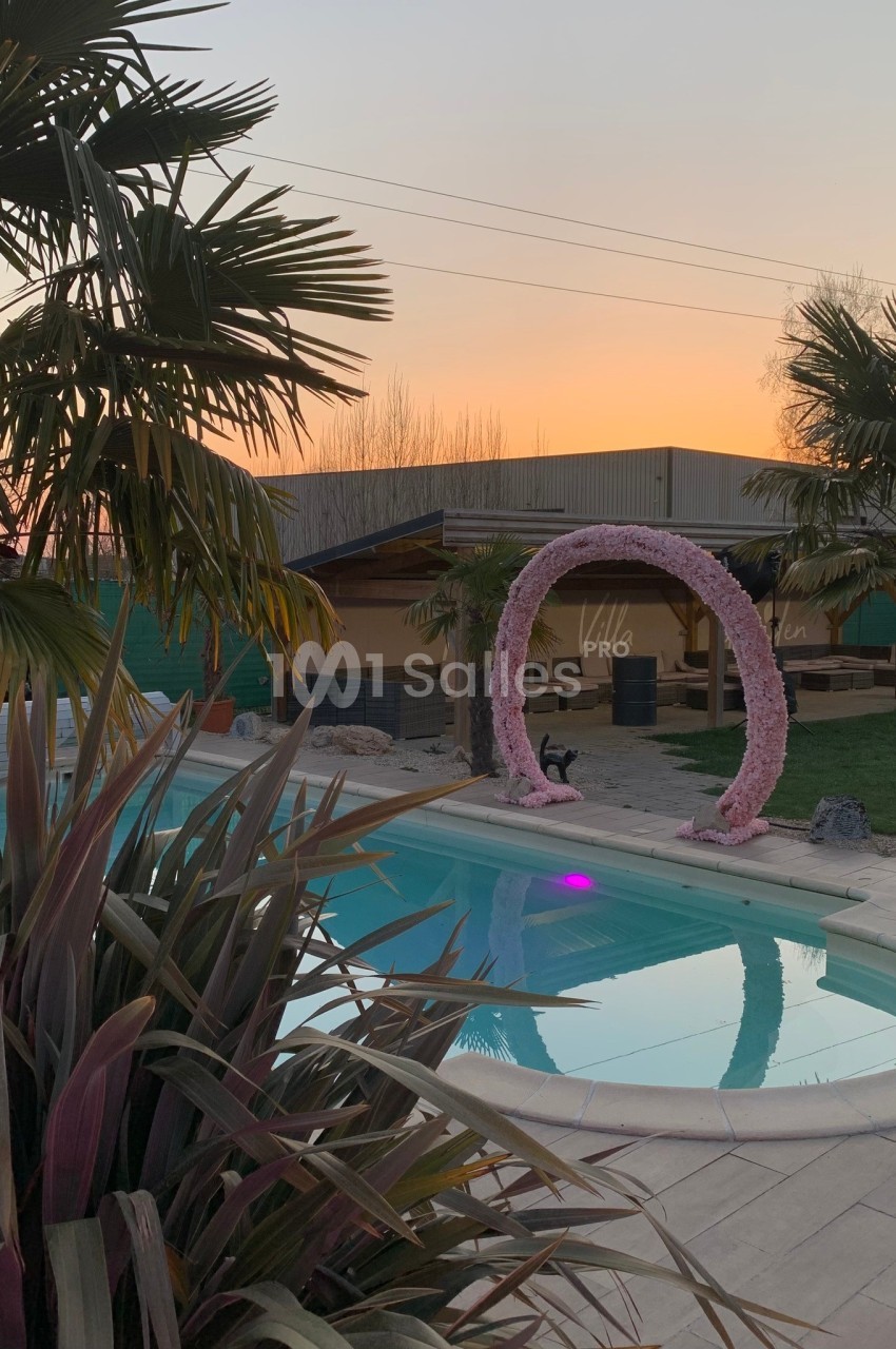 Piscine extérieure entourée de palmiers, avec une arche décorative et un coucher de soleil en arrière-plan.