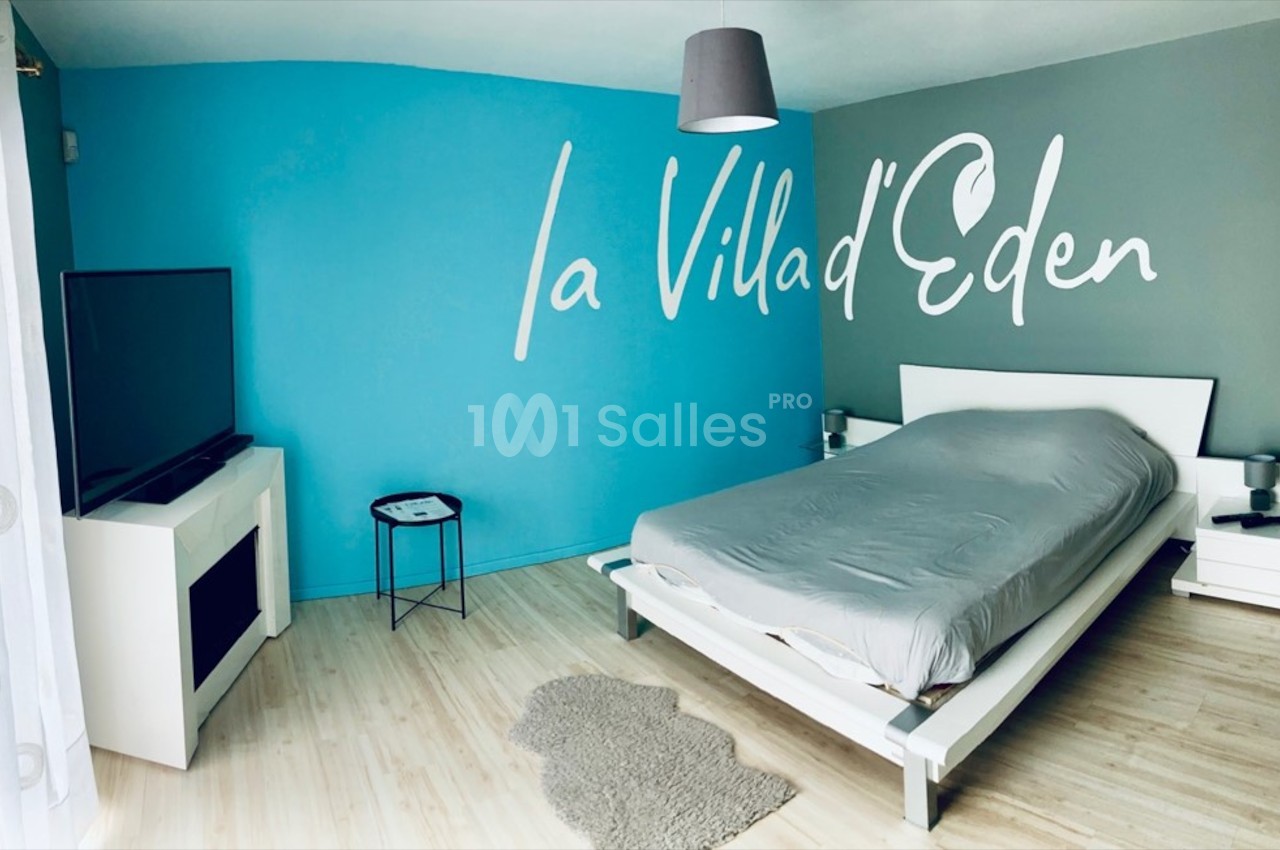 Chambre moderne avec un mur bleu, lit double, téléviseur, cheminée décorative et inscription ’La Villa d'Eden’.