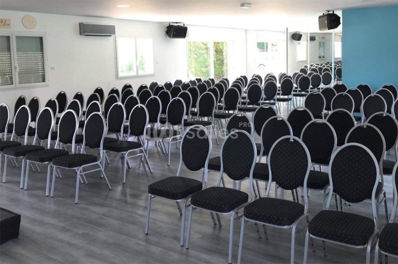 Salle lumineuse avec des rangées de chaises noires alignées sur un sol en parquet gris, prête pour un événement.
