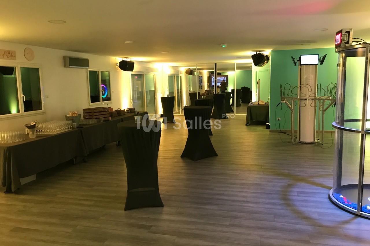 Salle lumineuse aménagée pour un événement, avec tables hautes, buffet et cabine photo dans un coin.