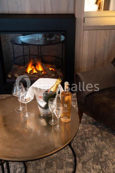 Bouteille de champagne, vin blanc et verres sur une table basse devant une cheminée allumée.