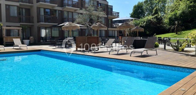 Piscine extérieure entourée de transats, parasols en paille et bâtiments modernes, avec jardin en arrière-plan.