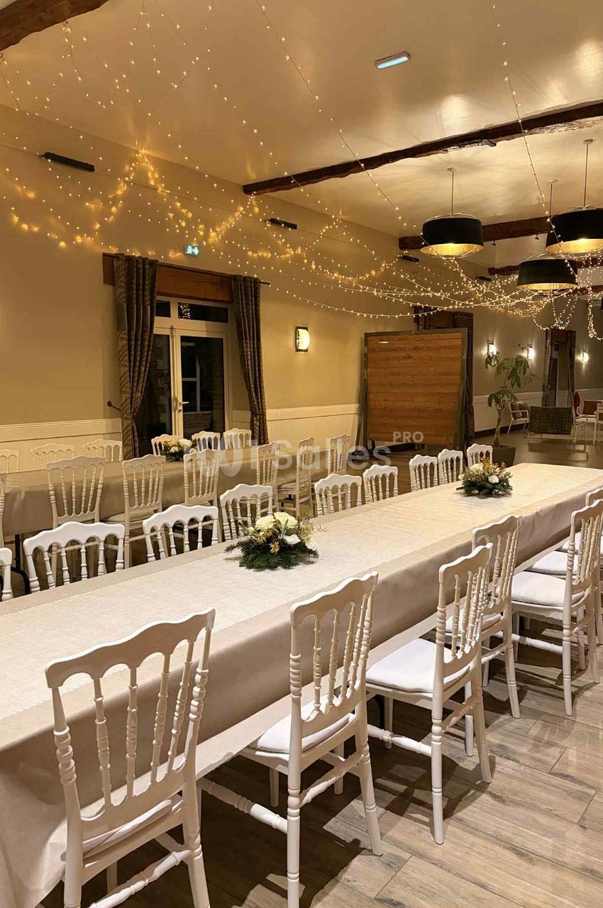 Salle décorée avec des guirlandes lumineuses, tables longues dressées avec nappes blanches et chaises alignées.
