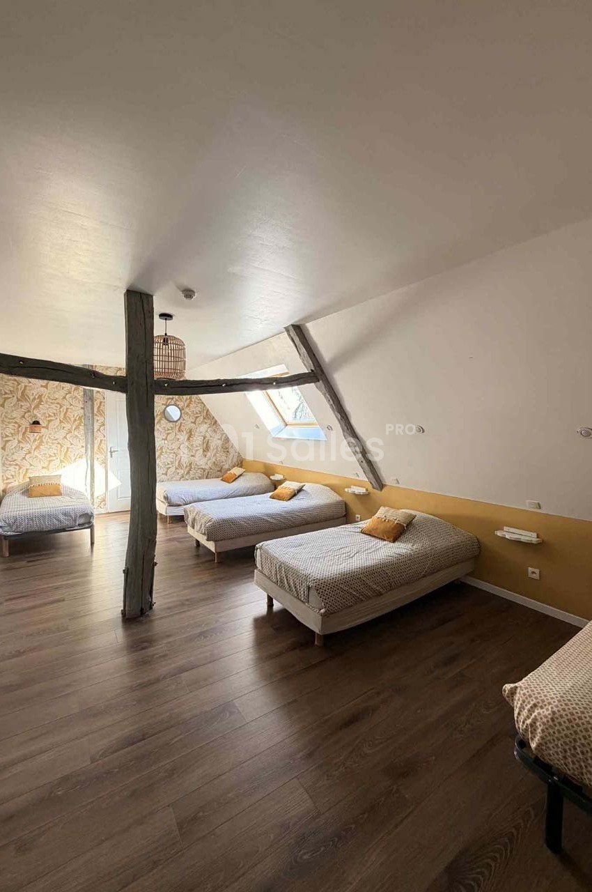 Chambre mansardée avec quatre lits simples, poutres apparentes, parquet et lumière naturelle provenant d'une fenêtre.