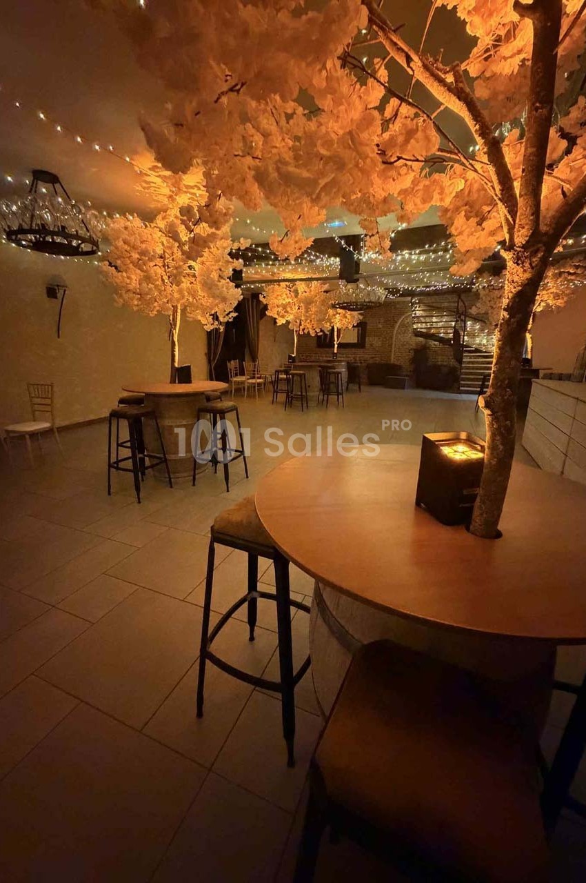 Salle décorée avec des arbres lumineux, des tables hautes et des guirlandes suspendues créant une ambiance chaleureuse.