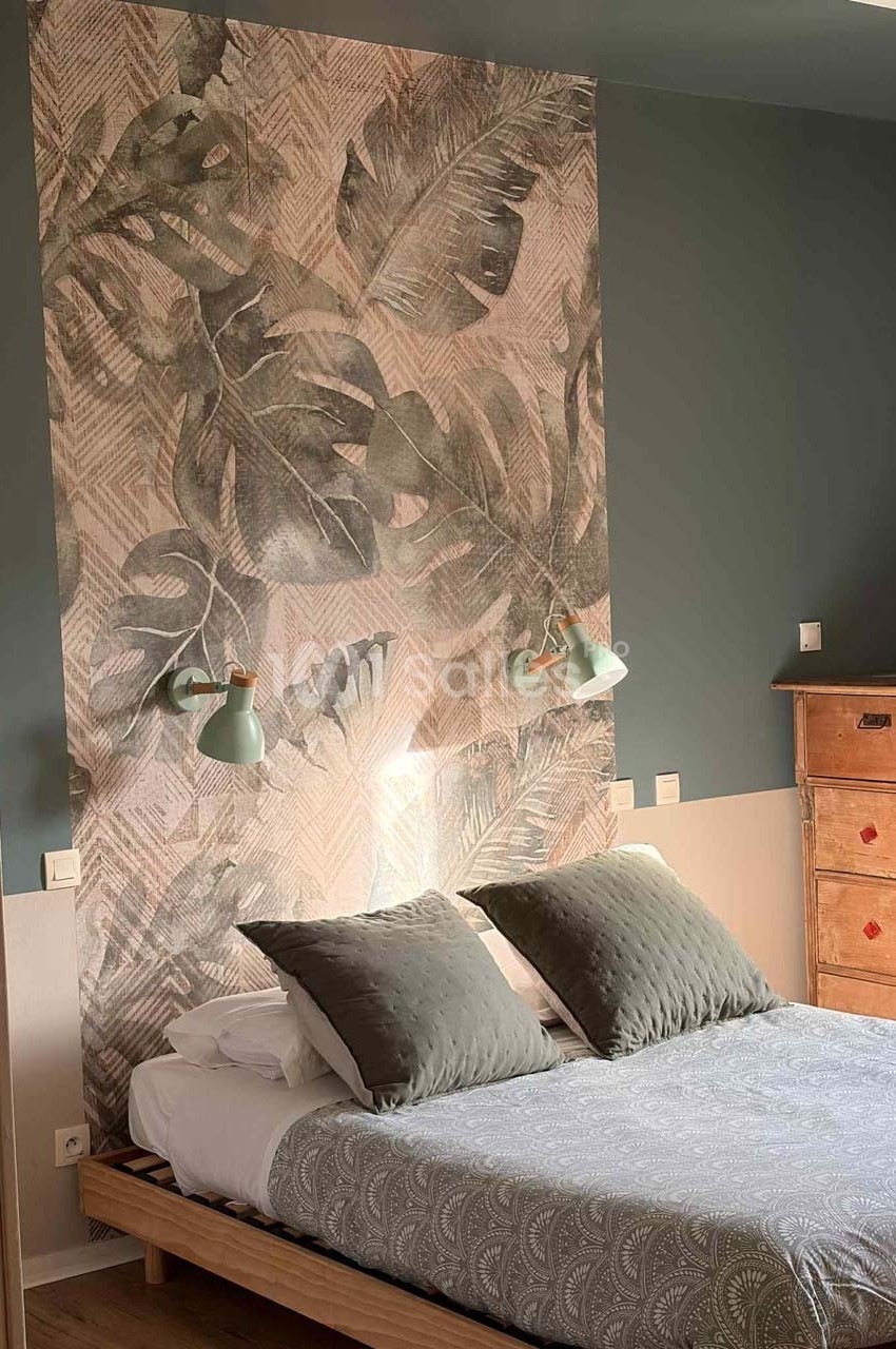Lit double avec coussins gris, tête de lit décorée de motifs végétaux, éclairée par deux appliques murales vertes.