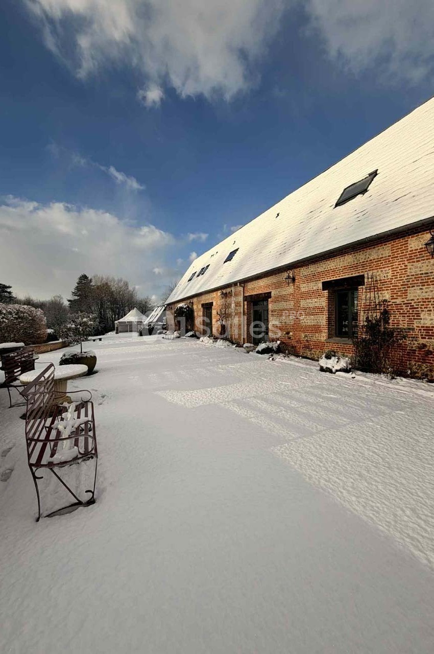 Cour d'une maison en briques sous la neige, avec bancs métalliques et ciel partiellement nuageux.