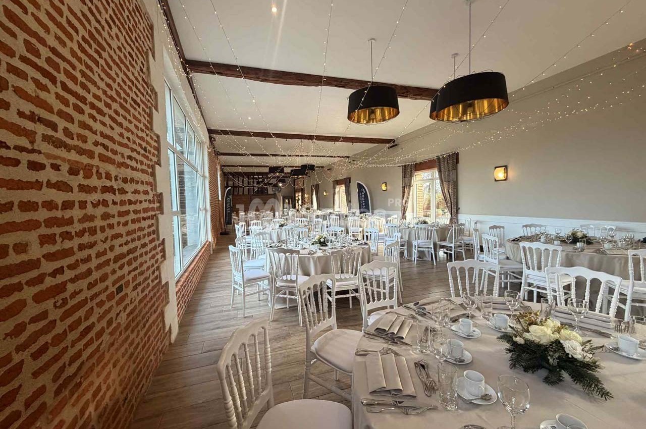 Salle de réception décorée avec des tables rondes dressées, chaises blanches et guirlandes lumineuses suspendues.