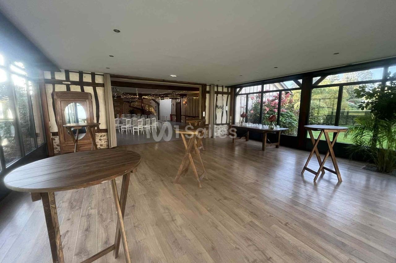 Salle lumineuse avec parquet, tables hautes en bois, grandes baies vitrées et vue sur un jardin fleuri.