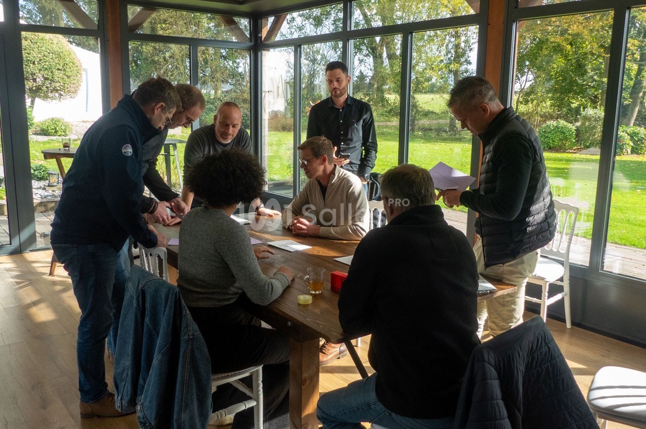 Un groupe de personnes discute autour d'une table en bois dans une pièce lumineuse avec vue sur un jardin.
