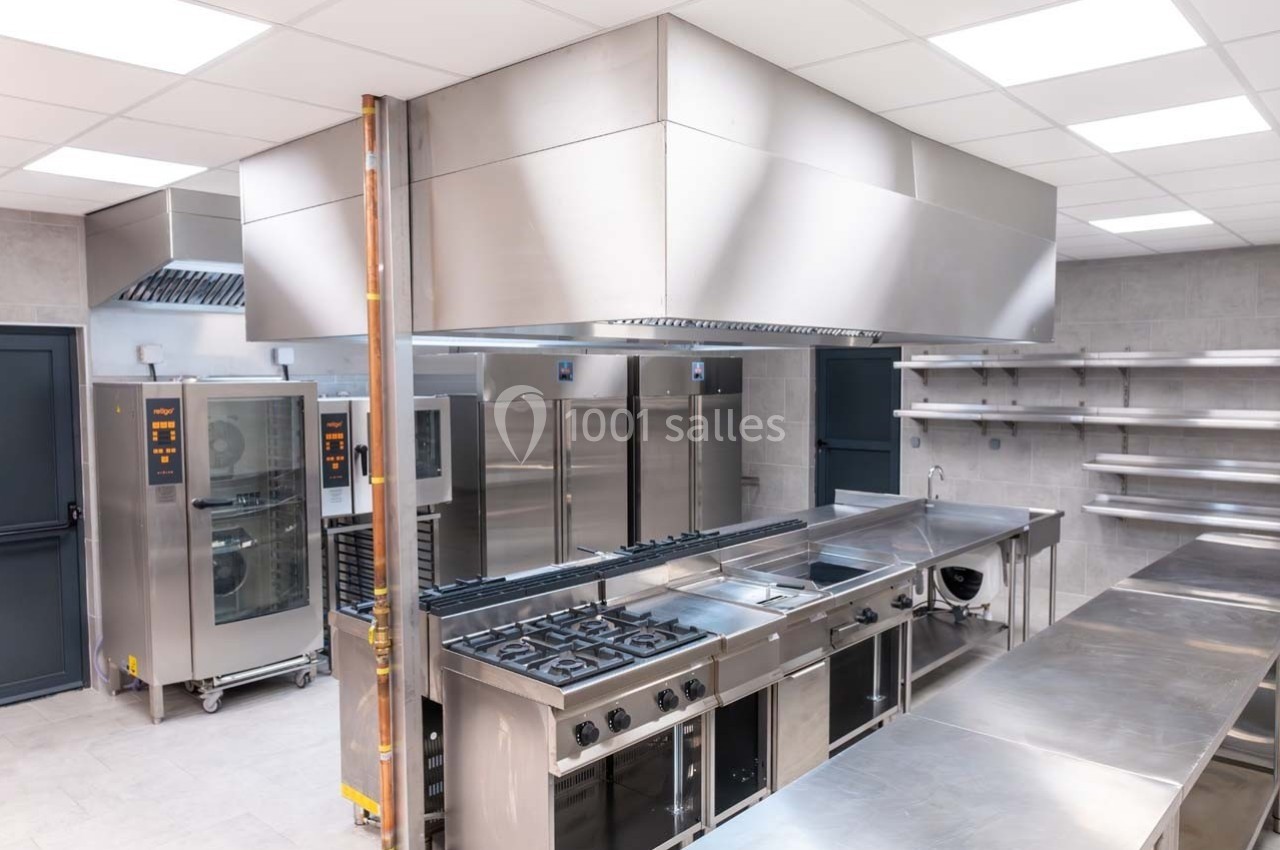 Cuisine professionnelle équipée avec plans de travail en inox, fours, plaques de cuisson et étagères murales.