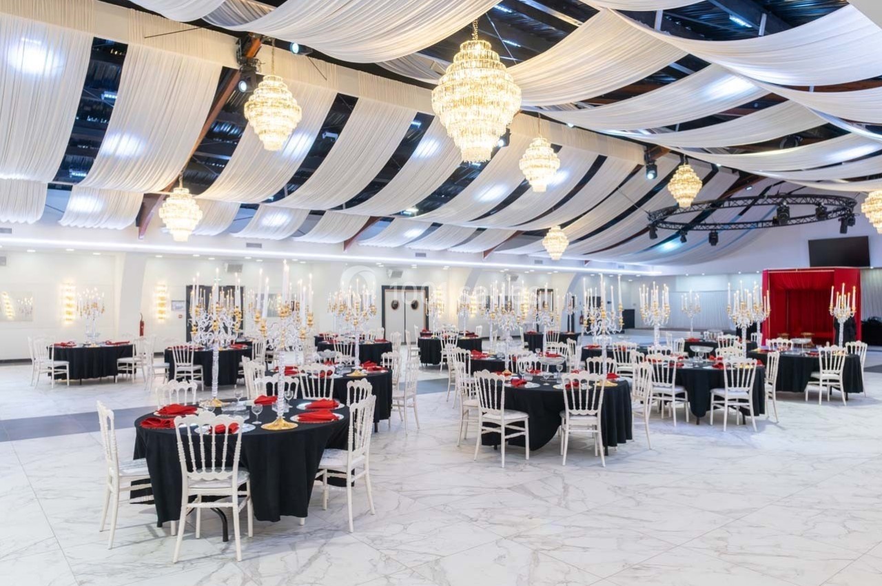 Salle de réception élégante avec tables dressées, nappes noires, chaises blanches et lustres suspendus sous des drapés…